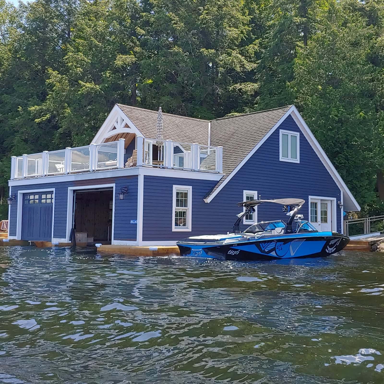 Navy blue boathouse with white trim, two slip doors at water level and a glass-railed upper deck, a boat moored alongside