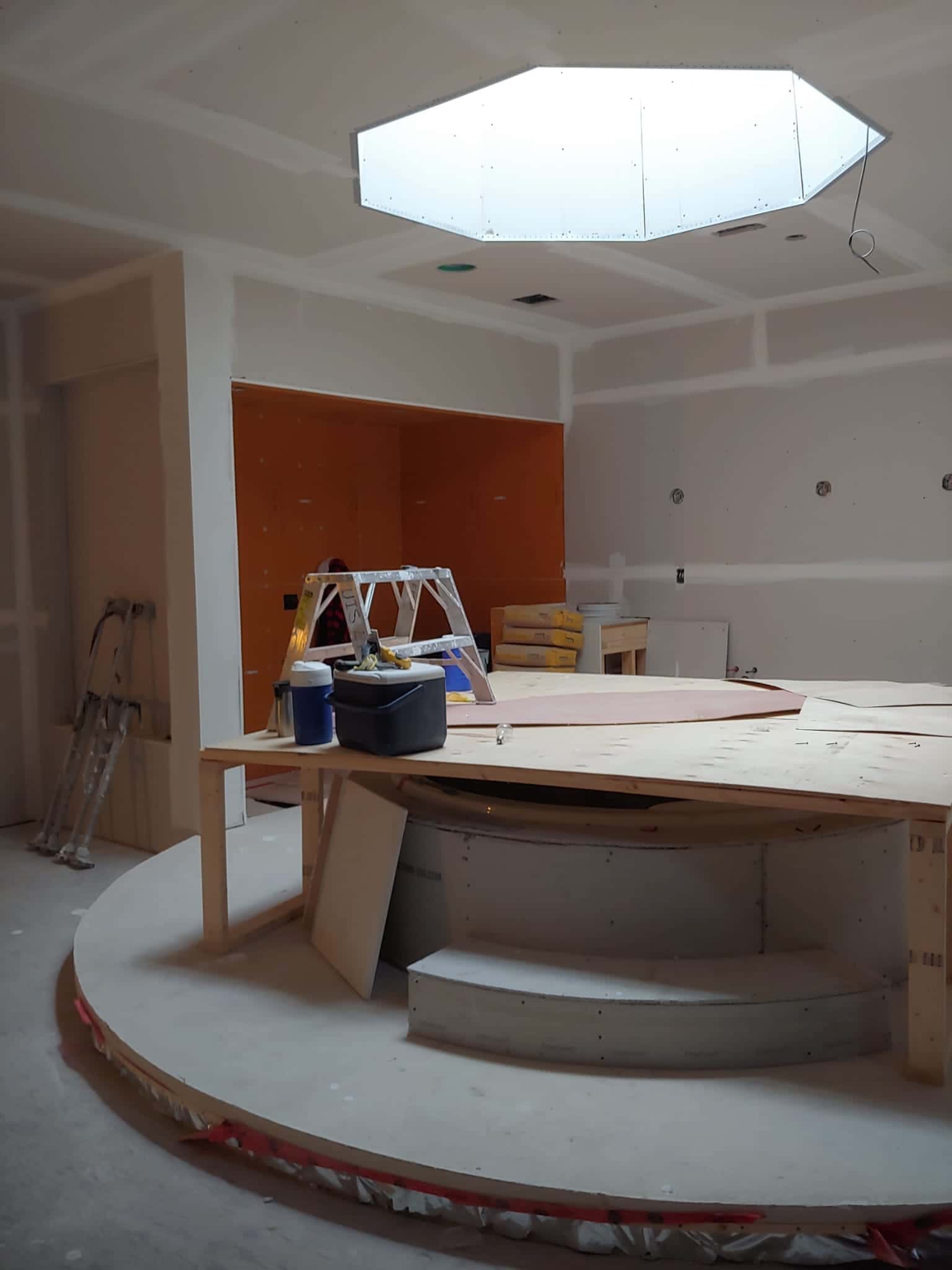 Interior at the drywall stage with an octagonal skylight well overhead, a coffered ceiling and a curved raised platform under plywood protection