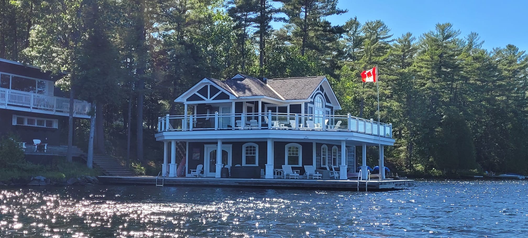 Two-storey boathouse with white columns and a glass-railed upper deck over open water, a Canadian flag flying beside it
